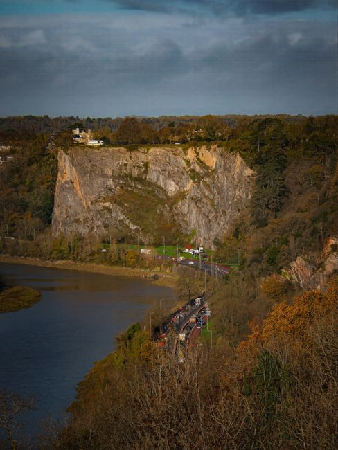 A scenic landscape featuring a large rocky cliff with a flat, grassy top, situated behind a winding river and dense trees displaying autumn foliage. In the distance, a small building is visible on the
