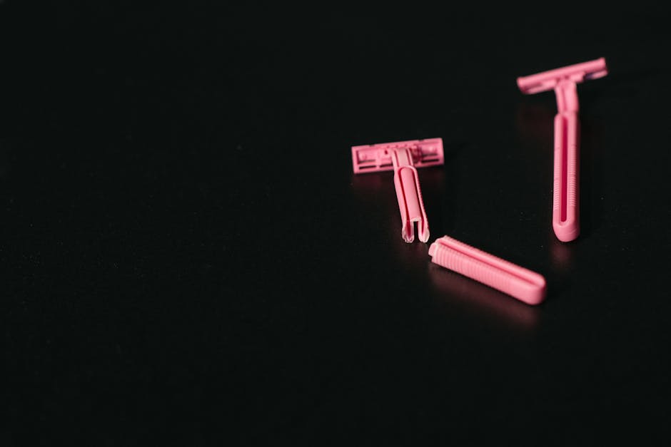 Three pink disposable razors with plastic handles and safety heads are placed on a black surface. The razors are arranged in a scattered manner, with one lying flat and the other two slightly angled. The dark background provides a high contrast, making the razors stand out clearly. The image captures the details of the razors' design, including the textured grip areas and the protective plastic covers over the blades. This simple, close-up view focuses on the razors' materials and form, without any additional objects or background distractions. The setting appears neutral and professional, suitable for topics related to personal care or hygiene products, and indirectly supporting content about packing or handling bathroom items during house moves in the context of professional removals by Man with Van Ratcliff.