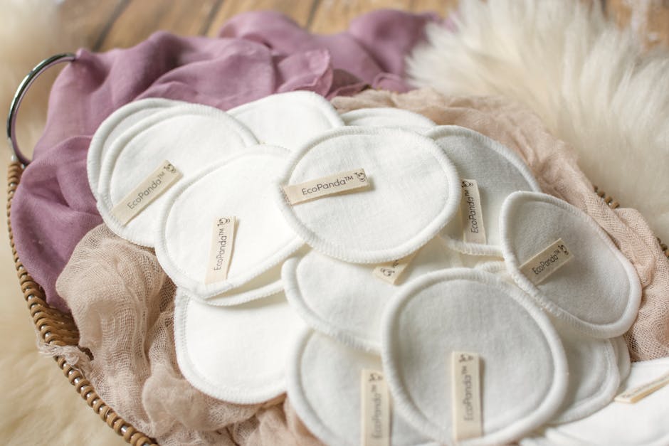 A woven basket filled with multiple white reusable eco-friendly makeup remover pads, each with a beige label reading 'EcoPanda,' resting on soft, folded fabrics in mauve, pink, and beige tones. The basket is placed on a light-colored, fluffy surface, likely a rug or blanket, with natural lighting highlighting the textures of the fabrics and pads. In the background, a glimpse of a white, fluffy object suggests a cozy, indoor setting. This scene illustrates the packing process for eco-conscious home relocation or household item transport, where reusable cleaning accessories are gathered for moving or disposal. The arrangement emphasizes attention to packing details and environmentally friendly practices, aligning with furniture transport and packing services provided by Man with Van Ratcliff, supporting efficient and responsible furniture transport in house removals.