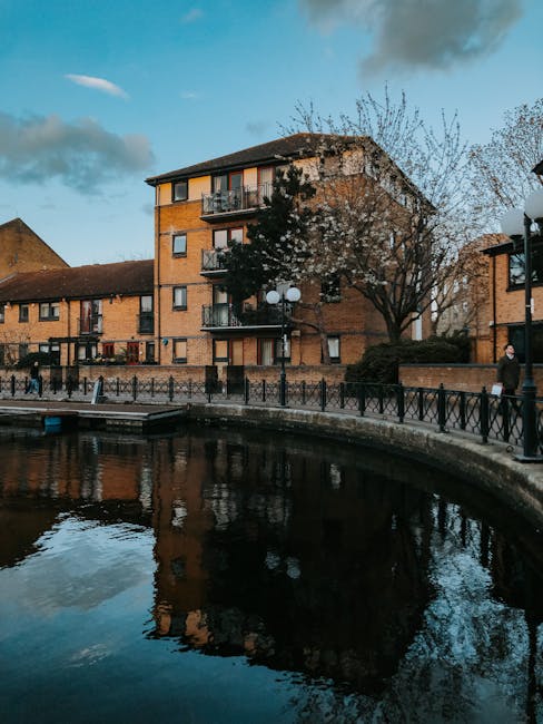 A multi-storey residential building made of brick with several balconies and windows, situated beside a canal with calm, reflective water. The building is surrounded by leafless trees and a paved walkway with metal railings, where a few pedestrians are visible walking alongside the water. A street lamp with multiple round white lanterns is positioned near the building entrance, and the sky is partly cloudy with soft, natural light. This scene captures an urban setting typical of a house relocation area near Limehouse Basin, illustrating the environment where Man with Van Ratcliff provides house removals and furniture transport services, including packing and loading processes surrounding the property and nearby access points.