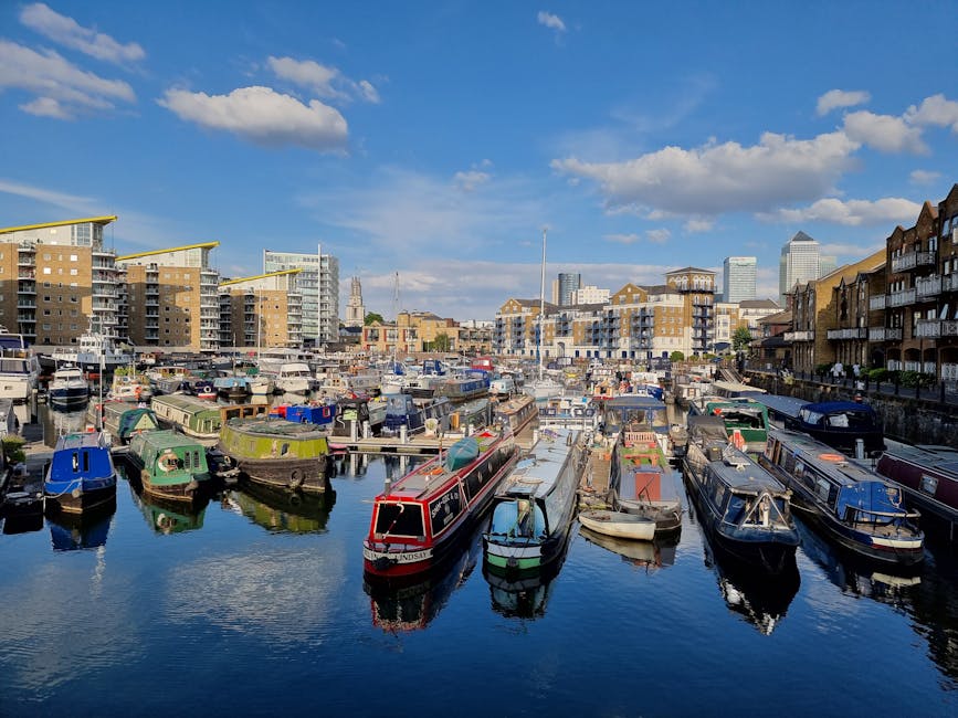The image shows a multi-story residential building with modern architectural design, featuring curved balconies, large glass windows, and a yellow brick facade. In the foreground, several boats are moored along the water's edge, with one boat prominently displaying the name 'LEONE' on its side, indicating a marina or dock area near Limehouse Basin. The boats vary in size and design, with some covered by tarps and others exposed to the open air. Lush green trees are situated at the base of the building, partially obscuring the lower levels and providing a natural contrast to the urban environment. The scene is illuminated by daylight with a background of a partly cloudy sky, suggesting clear weather conditions. The composition captures the proximity of residential living spaces to the waterfront, relevant to local house removals, furniture transport, and moving logistics provided by Man with Van Ratcliff, particularly for short-notice relocations near Ratcliff.