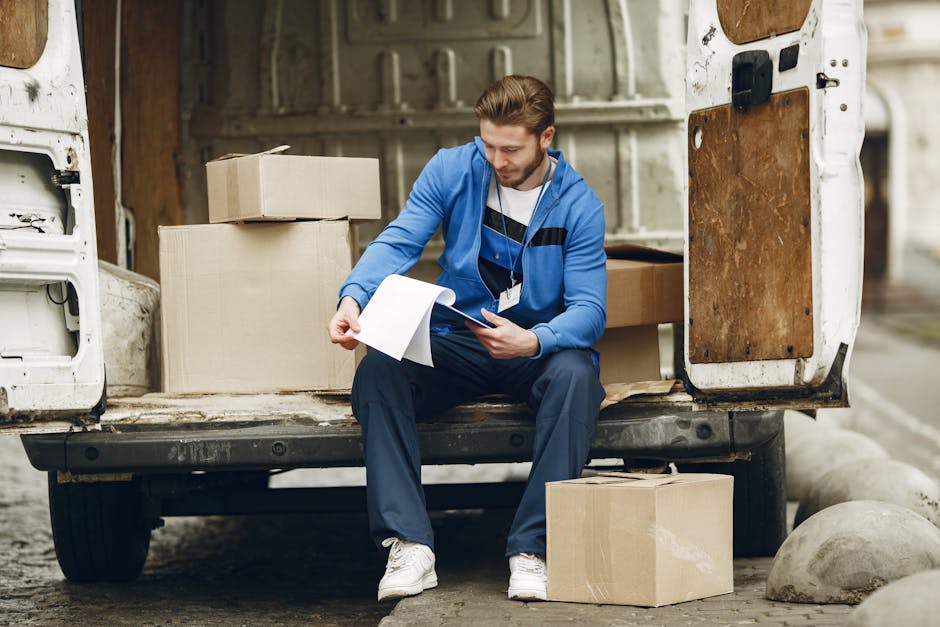 A man with light brown hair, wearing a blue zip-up hoodie, dark blue work pants, and white sneakers, is seated on the edge of an open white van during a home relocation process. He is holding a clipbo