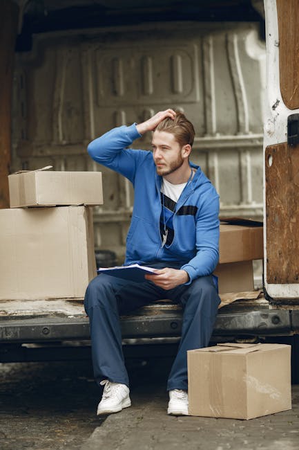 A man with light brown hair, wearing a blue zip-up hoodie, dark blue work pants, and white sneakers, is seated on the edge of an open white van during a home relocation process. He is holding a clipbo