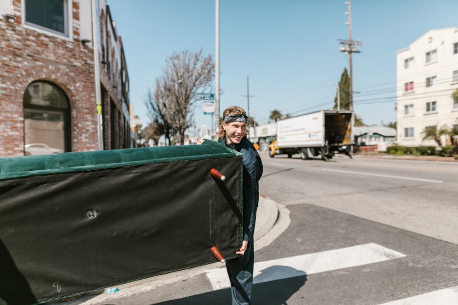 A man with a headband smiling as he carries a large, black padded moving blanket over his shoulder during a home relocation process outside a residential building. The blanket, featuring orange handles, is used to protect furniture during transport. He stands on the sidewalk near a street intersection, with a busy road visible in the background, including a white delivery truck and other vehicles. The setting is outdoors in a city area with brick and white multi-storey buildings, trees, and utility poles visible in the distance. The scene is well-lit with natural daylight, illustrating the loading process involved in a local house removal carried out by Man with Van Ratcliff, who manage furniture transport and packing during moving services.