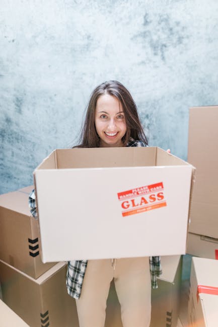 A woman with long brown hair smiling at the camera while holding a large empty cardboard box labeled 'Glass' during a home relocation process. She is standing inside a room with a light blue, textured wall and surrounded by other packed cardboard boxes of various sizes, some sealed with packing tape. The boxes are made of brown corrugated cardboard, and some have black arrow symbols indicating orientation. The woman is wearing a casual plaid shirt and light trousers, suggesting she is involved in packing or moving activities as part of a local removals service by Man with Van Ratcliff. The scene captures the loading or packing stage of furniture transport and house removals, with a focus on organized packing materials and moving preparation.