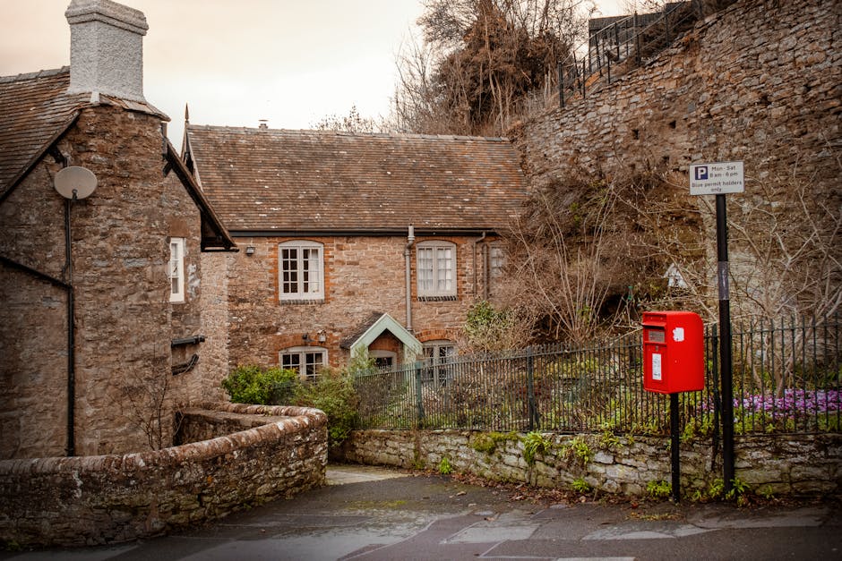 A residential street scene featuring an old stone house with a pitched roof and white-framed windows, partially obscured by leafless shrubbery and small plants. In the foreground, a black metal fence runs along a low stone wall, separating the property from the paved pavement. To the right, a red postbox is mounted on a black metal pole near a sign indicating parking restrictions for Monday to Saturday from 8 am to 6 pm, with a note that blue permit holders only are allowed. The background includes a hillside with dry grass and a stone retaining wall topped with a black metal fence. The scene is captured during daylight with overcast or diffused lighting, and there is no visible activity or moving objects, just the exterior features of a typical home, relevant to property access and infrastructure considerations for home relocation, furniture transport, or packing and moving processes as handled by Man with Van Ratcliff.