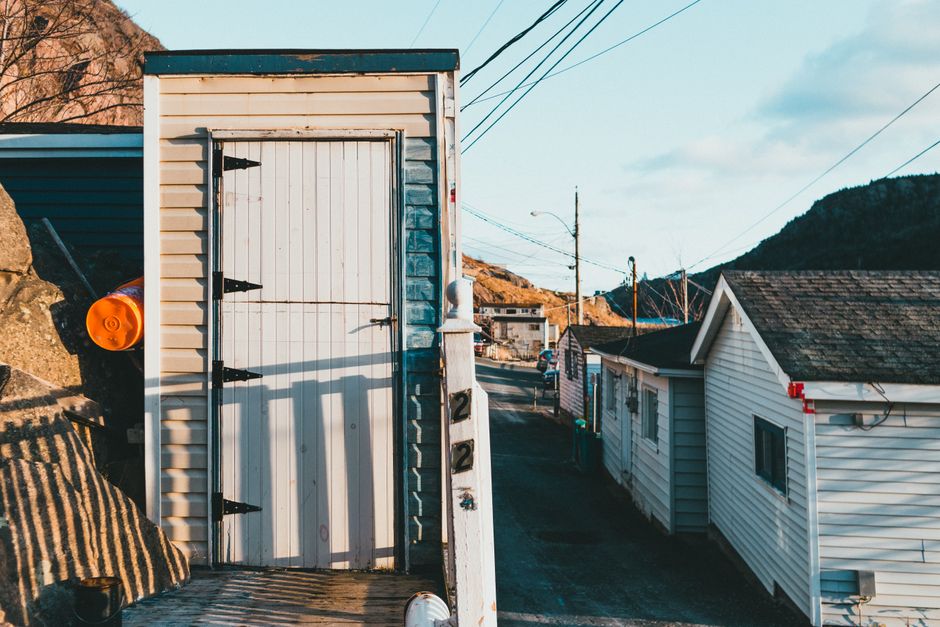 A white shed with horizontal siding and a vertical wooden door secured with black hinges and a latch, situated on a slightly elevated outdoor area. In front of the shed, there are several cardboard boxes and packing materials, indicating a home relocation process. To the left, an orange safety light is mounted on the shed's exterior, and on the ground, a striped fabric moving blanket is partially visible. The surrounding environment includes a paved driveway leading down the street lined with residential houses, one of which is a small white house with a sloped roof, and a utility pole with overhead wires extends into the sky. The scene is lit by natural daylight, with clear weather and distant hills visible in the background. This setting reflects typical furniture transport and packing activities involved in house removals, as managed by companies like Man with Van Ratcliff.