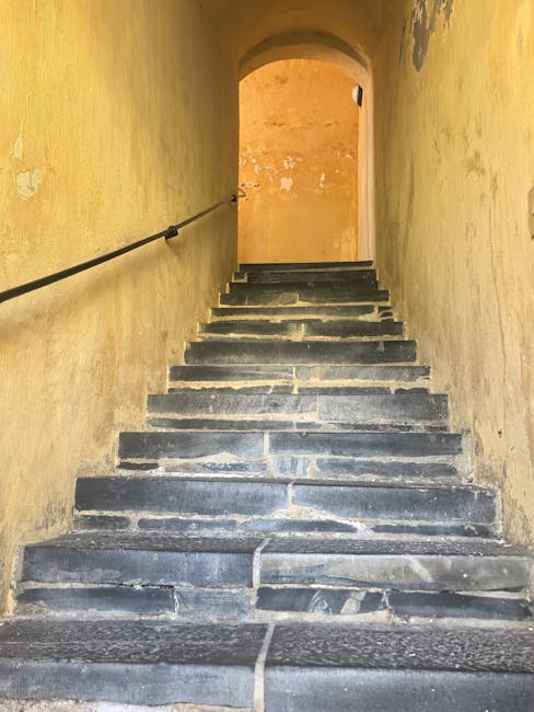 A narrow, steep stone staircase inside a historic building with worn, uneven steps made of dark stone and visible signs of use. The staircase is enclosed by yellowish, textured walls that show some minor peeling paint and age, and a black metal handrail runs along the left side, providing support for individuals ascending or descending. At the top of the stairs, there is an arched doorway leading into another room or corridor with a warm, golden-toned wall. This setting is part of a property where home relocation or moving services, such as those provided by Man with Van Ratcliff, may involve navigating tight and challenging staircases during furniture transport or packing and moving processes, especially in buildings with historical features. The lighting inside is natural or softly diffused, illuminating the stairs and highlighting their worn surface, emphasizing the need for careful handling during a house removal.
