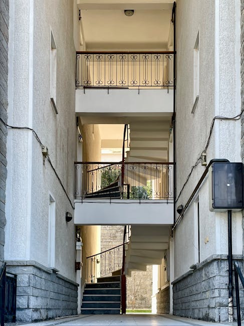The image shows an outdoor stairwell in a residential building, featuring three flights of cream-colored concrete stairs connected by landings with black wrought iron railings. The stairwell is enclosed by plain white walls with small rectangular windows on each side; some are open, allowing natural light into the space. On the left wall, there are visible black cables running along the surface. At the bottom, a dark grey mat covers the ground just outside the entrance, which leads to the street or courtyard area. The stairwell appears clean and well-maintained, with some greenery visible through the open yard beyond the lower landing. This setting is typical of urban residential buildings, where internal staircases facilitate access to upper floors. Man with Van Ratcliff utilizes such staircases during house removals and furniture transport, carefully maneuvering items through narrow indoor spaces as part of their house relocation services, ensuring efficient and safe loading and unloading processes.