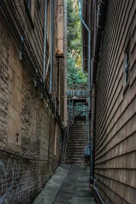 A narrow, steep stone staircase inside a historic building with worn, uneven steps made of dark stone and visible signs of use. The staircase is enclosed by yellowish, textured walls that show some minor peeling paint and age, and a black metal handrail runs along the left side, providing support for individuals ascending or descending. At the top of the stairs, there is an arched doorway leading into another room or corridor with a warm, golden-toned wall. This setting is part of a property where home relocation or moving services, such as those provided by Man with Van Ratcliff, may involve navigating tight and challenging staircases during furniture transport or packing and moving processes, especially in buildings with historical features. The lighting inside is natural or softly diffused, illuminating the stairs and highlighting their worn surface, emphasizing the need for careful handling during a house removal.