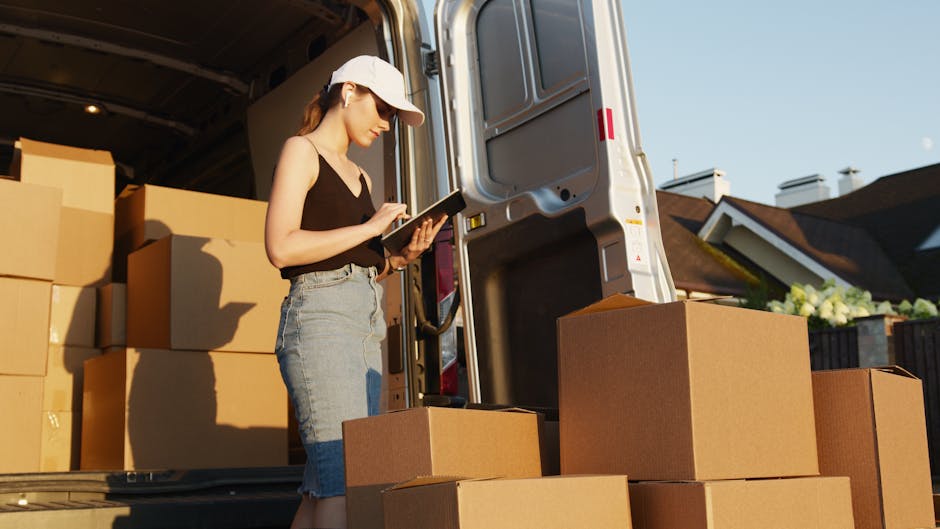 A woman wearing a white cap, black tank top, and light blue denim skirt stands next to the open rear door of a grey moving van, inspecting or taking notes on a tablet device. She is surrounded by neatly stacked cardboard boxes of various sizes, some inside the van and others placed on the pavement outside. The boxes are sealed with packing tape and likely contain household items prepared for a home relocation or furniture transport. The interior of the van appears organized, with additional packing materials such as plastic wrap or blankets possibly used to protect furniture during loading. Behind her, residential houses with pitched roofs and chimneys are visible under a clear blue sky, indicating daytime. The scene captures the loading process involved in professional moving services, with emphasis on careful packing, documentation, and logistics coordination, as provided by Man with Van Ratcliff, in accordance with same-day move requirements.
