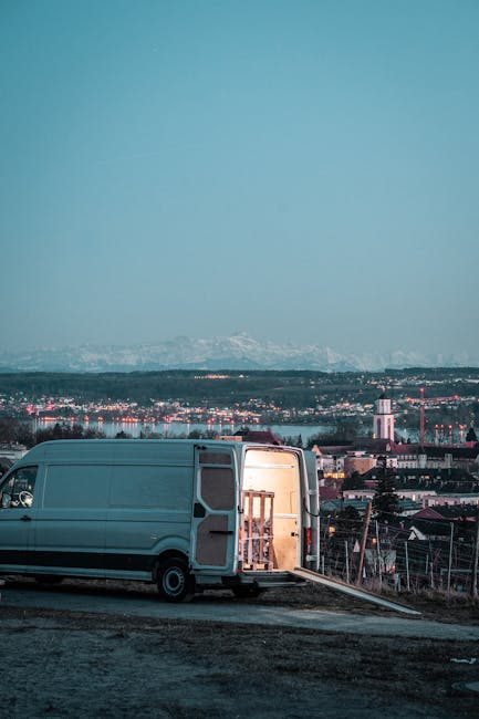 A large, white moving van parked on an outdoor surface, with its rear doors open revealing a wooden furniture piece and packing materials inside, including cardboard boxes and plastic wrap. The van is positioned near the edge of a site with a slight slope, and a ramp extends from the ground into the vehicle's interior to facilitate loading or unloading. In the background, there is a panoramic view of a cityscape with buildings, industrial structures, and distant snow-capped mountains under a clear sky during twilight or early evening. This scene depicts a home relocation or furniture transport process associated with professional removals services, such as those offered by Man with Van Ratcliff.