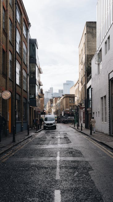 A narrow urban street scene during daylight showing modern brick and concrete buildings lining both sides of the road, with a white van parked near the sidewalk and several pedestrians walking along the pavement. The street surface appears wet, suggesting recent rain, and the sky is overcast. In the background, taller buildings and a hint of the city skyline are visible. The scene captures a typical setting for house removals or furniture transport in central London, illustrating an accessible route for a home relocation or moving services by Man with Van Ratcliff. The image emphasizes an urban environment suitable for logistical planning of packing and loading processes for removals within the E1 postcode area, with the street’s narrowness highlighting the importance of optimal van routes for efficient house moves.