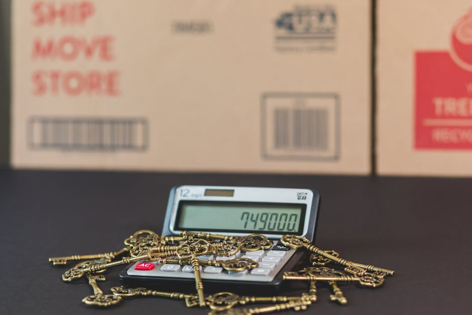 A calculator displaying the number 749,800 resting on a black surface, surrounded by a collection of gold-colored antique-style keys of various shapes and sizes. In the background, partially visible, are stacked cardboard boxes with red and blue printed labels, indicating packaging materials for a home relocation or moving process. The scene is well-lit, emphasizing the contrast between the keys, calculator, and boxes, representing aspects of packing and moving logistics related to furniture transport or house removals, with the presence of boxes and moving equipment aligning with professional moving services such as those offered by Man with Van Ratcliff.