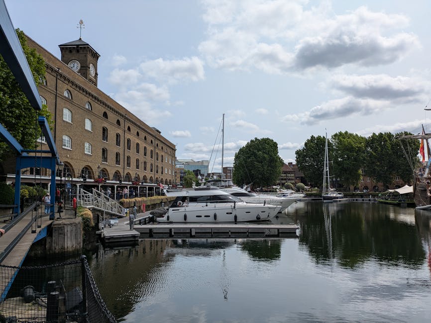 A view of a marina at the historic St Katharine Docks with several yachts and boats docked along the water, which reflects the cloudy sky above. On the left side of the image, there is a brick buildin