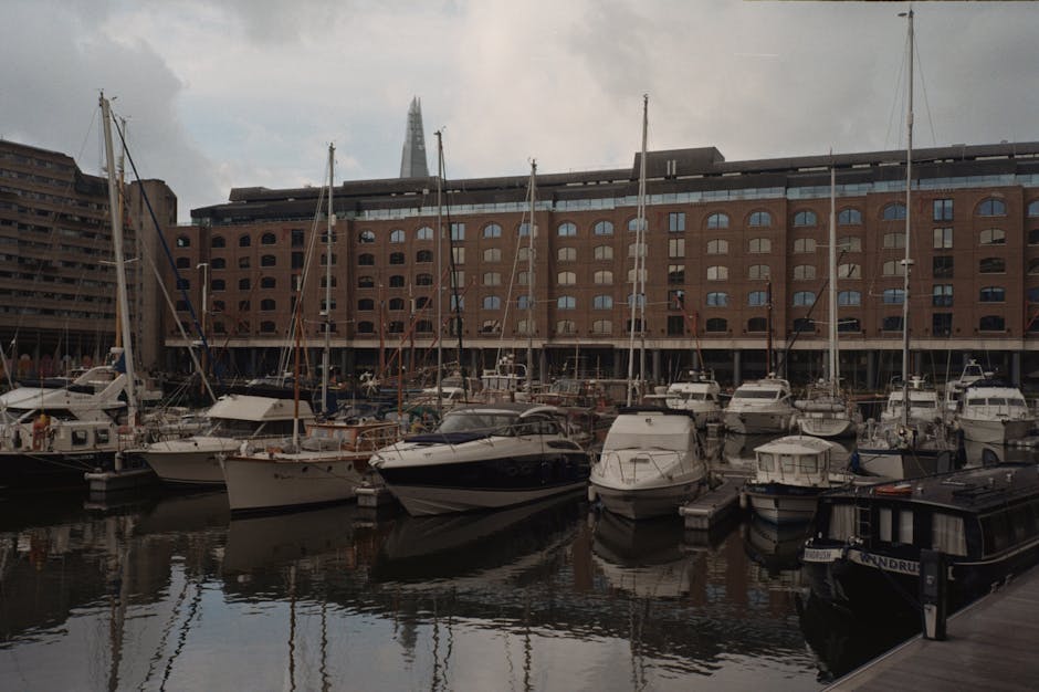 A view of a marina at the historic St Katharine Docks with several yachts and boats docked along the water, which reflects the cloudy sky above. On the left side of the image, there is a brick buildin