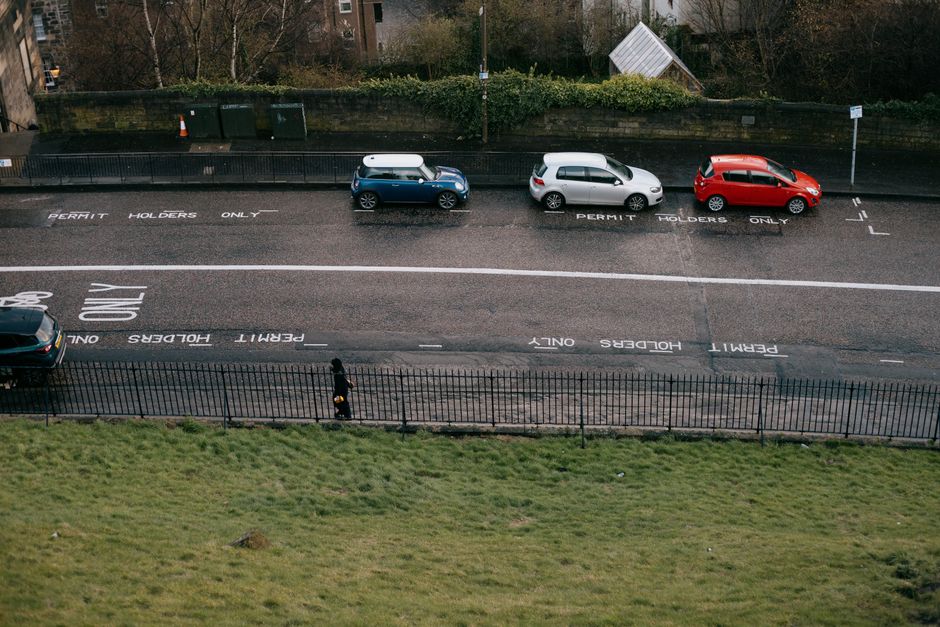 An aerial view of a city street showing a row of three parked cars, including a blue compact vehicle, a white hatchback, and a red hatchback, all aligned within designated parking bays marked with 'PERMIT HOLDERS ONLY' on the wet asphalt surface. The parking area is bordered by a curb and a green grassy strip with a metal fence separating the road from the park or green space below. The street appears to be part of a residential or urban area, with trees and bushes visible along the background. There are no visible moving or loading activities in this image, but it reflects typical parking arrangements associated with home relocation or furniture transport processes managed by professional removals services like Man with Van Ratcliff, who may assist with parking permits for efficient house moves in Tower Hamlets.