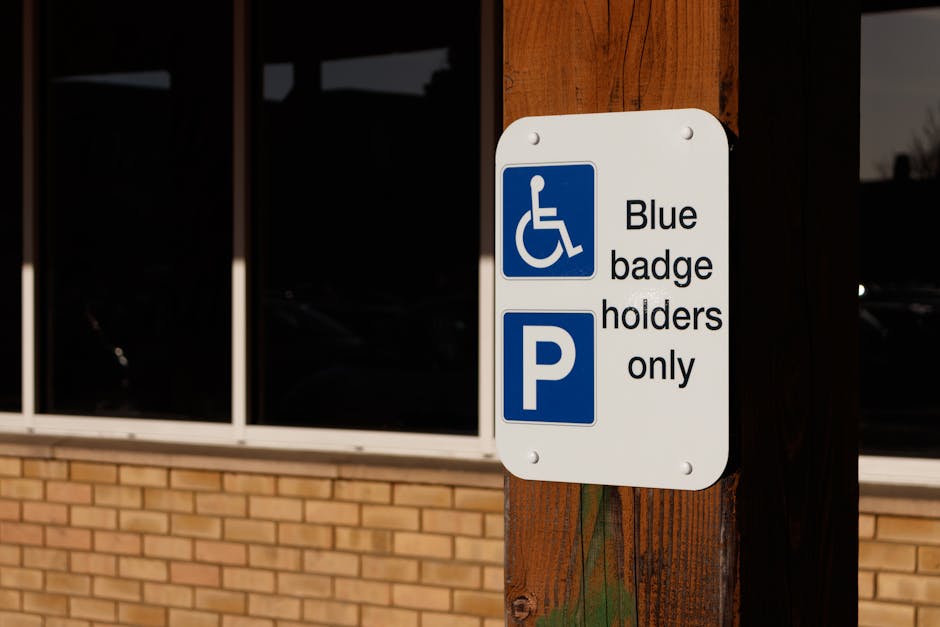 A white rectangular parking sign mounted on a wooden post outside a building with large glass windows and a brick lower wall. The sign displays a blue wheelchair symbol indicating accessibility for disabled badge holders, along with the text 'Blue badge holders only'. Below the wheelchair icon, there is a blue parking symbol with a 'P' and the service category is related to house removals and furniture transport. The sign is photographed in natural lighting, with reflections visible on the window glass, and the area appears to be designated for individuals with disabled parking permits, supporting the context of moving or relocation services provided by Man with Van Ratcliff.